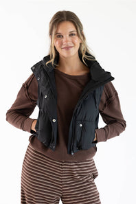 A woman modeling a black quilted puffer vest over a brown long-sleeve top and brown striped pants, photographed against a plain white studio background.