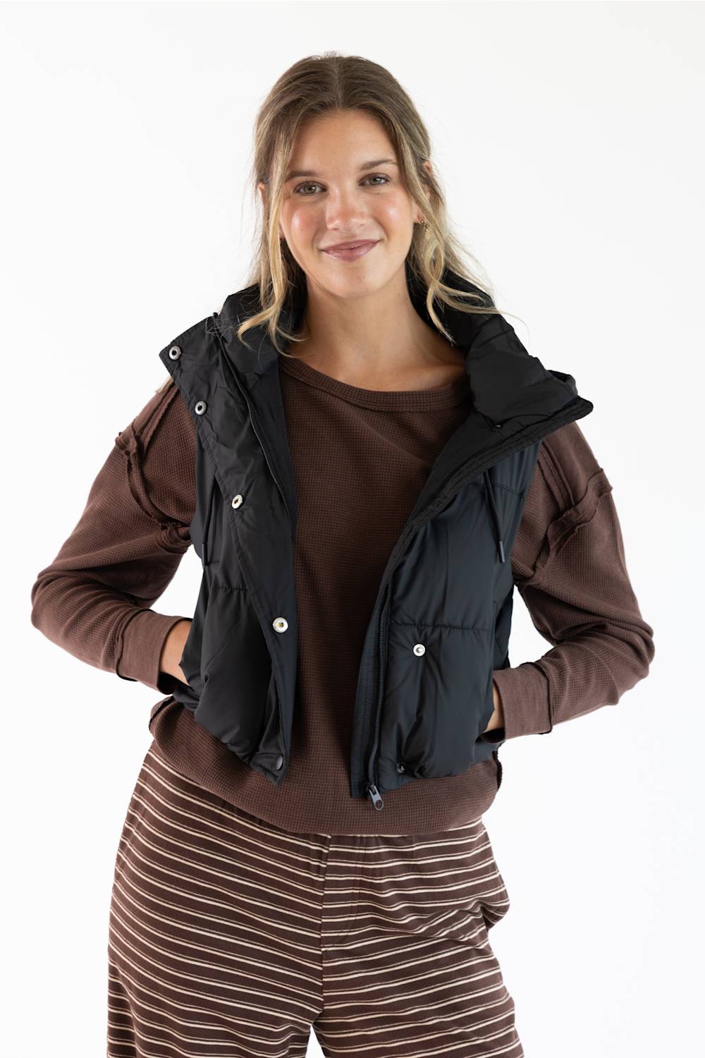 A woman modeling a black quilted puffer vest over a brown long-sleeve top and brown striped pants, photographed against a plain white studio background.
