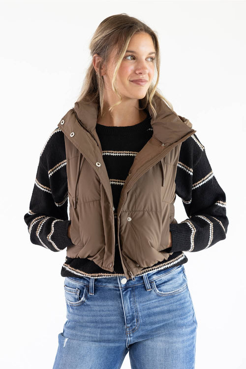 A woman wearing a brown sleeveless puffer vest over a black and white striped knit sweater, paired with blue jeans, against a plain white background.