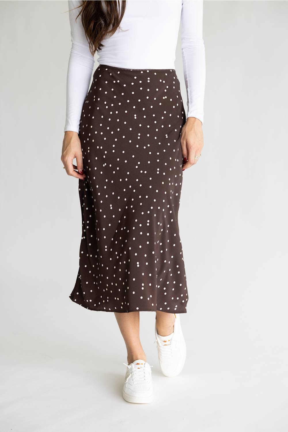 A woman wearing a brown midi skirt with a white polka-dot pattern, paired with a white long-sleeve top and white sneakers, shown from the waist down against a light gray studio background.