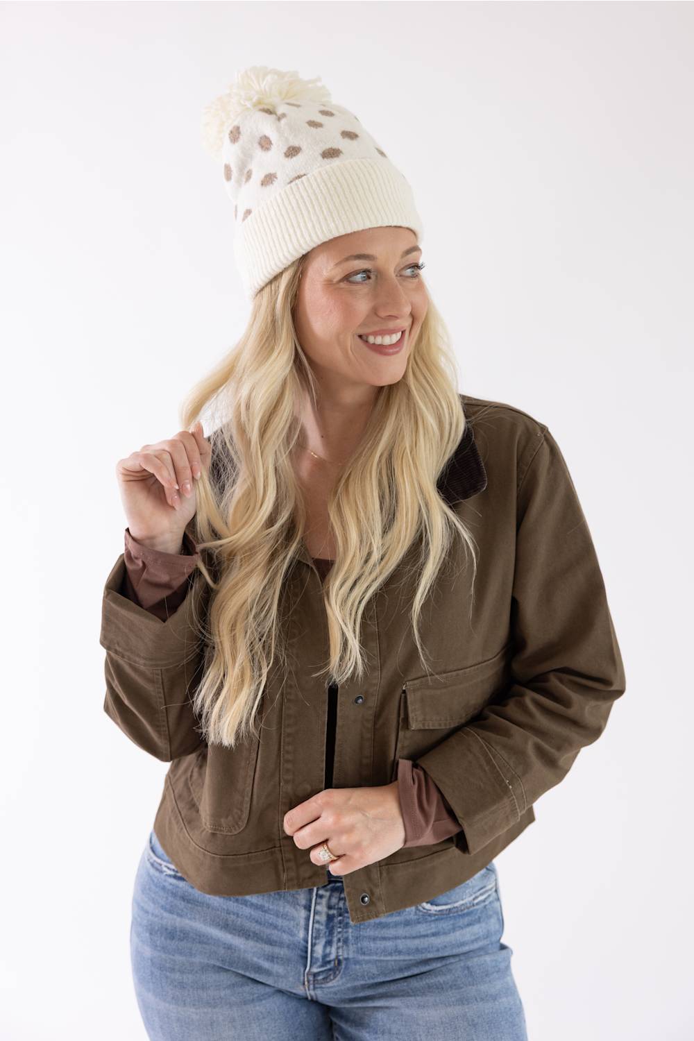 A woman wearing a brown utility jacket, cream knit beanie with brown spots, and blue jeans, smiling and posing against a plain white studio background.