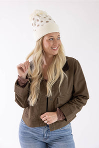 A woman wearing a brown utility jacket, cream knit beanie with brown spots, and blue jeans, smiling and posing against a plain white studio background.