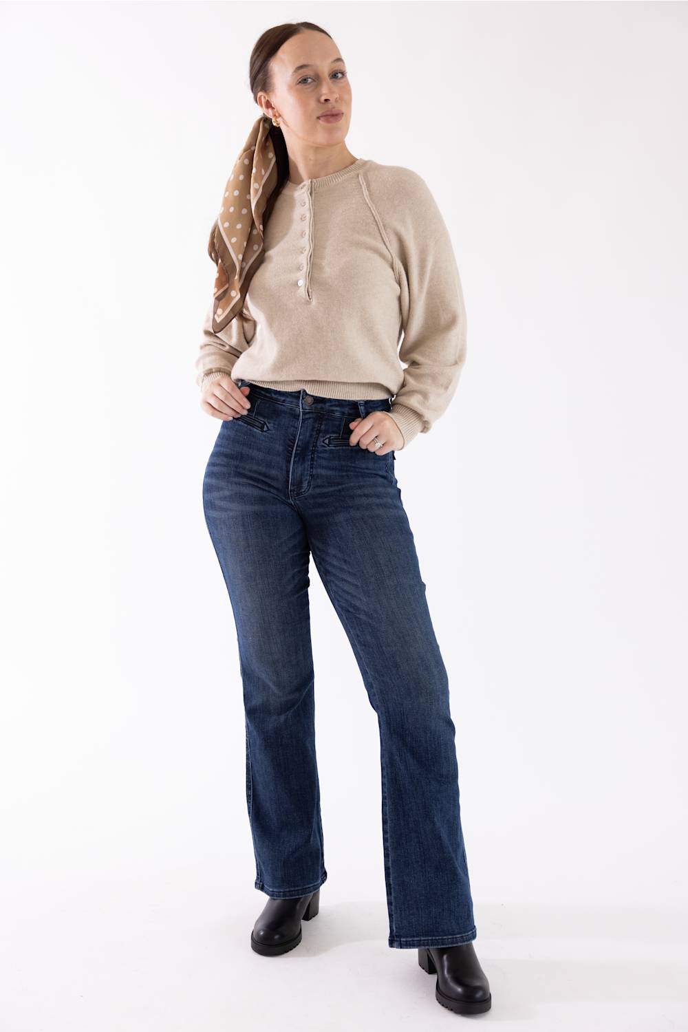 A woman modeling a beige knit sweater and dark blue straight-leg jeans against a plain white studio background.