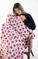 A woman sitting on a wooden chair, smiling as she wraps a light pink blanket with a red and black bow pattern around herself against a plain white background.