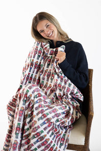 A smiling woman sits on a woven chair wrapped in a soft blanket featuring a repeating nutcracker soldier pattern on a white background.