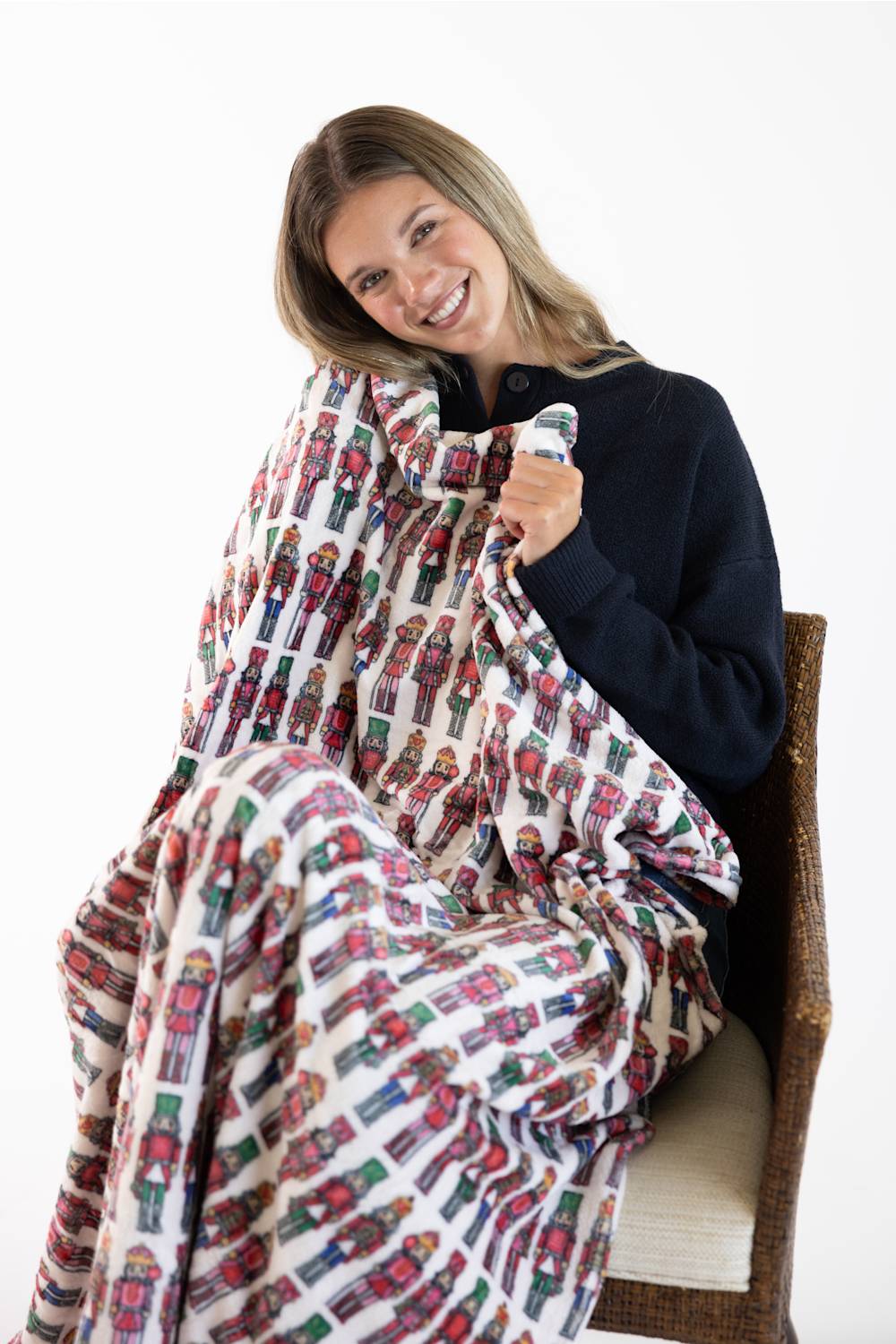 A smiling woman sits on a woven chair wrapped in a soft blanket featuring a repeating nutcracker soldier pattern on a white background.