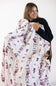 A woman holding up a white fleece blanket with a pink striped pattern and a whimsical print of pink bows, red stocking and Santa Clause, wearing a black long-sleeve top against a plain white background.