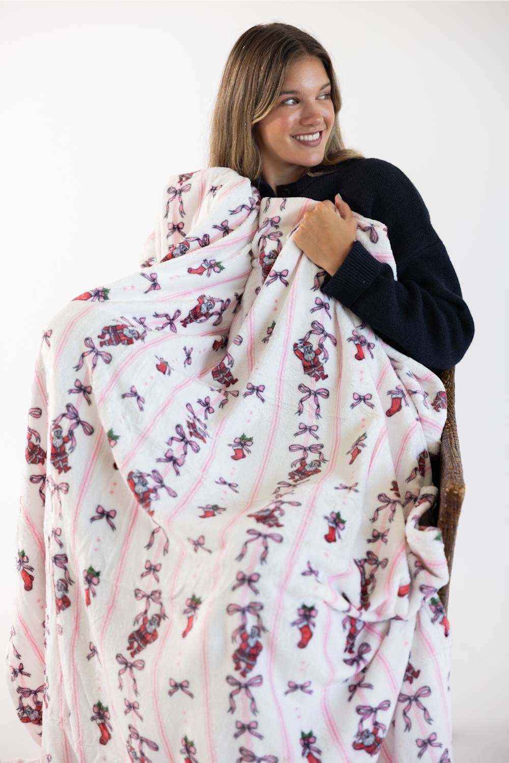 A woman holding up a white fleece blanket with a pink striped pattern and a whimsical print of pink bows, red stocking and Santa Clause, wearing a black long-sleeve top against a plain white background.