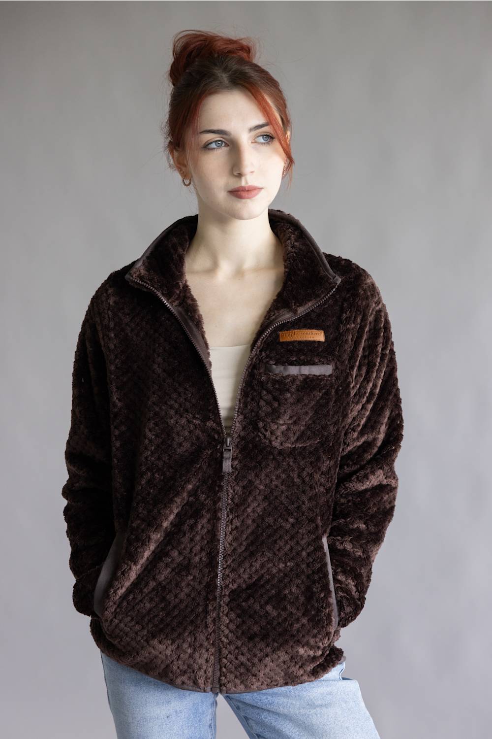 A woman with red hair in a bun wearing a dark brown textured zip-up fleece jacket, a cream top, and light blue jeans, standing against a neutral gray studio background.