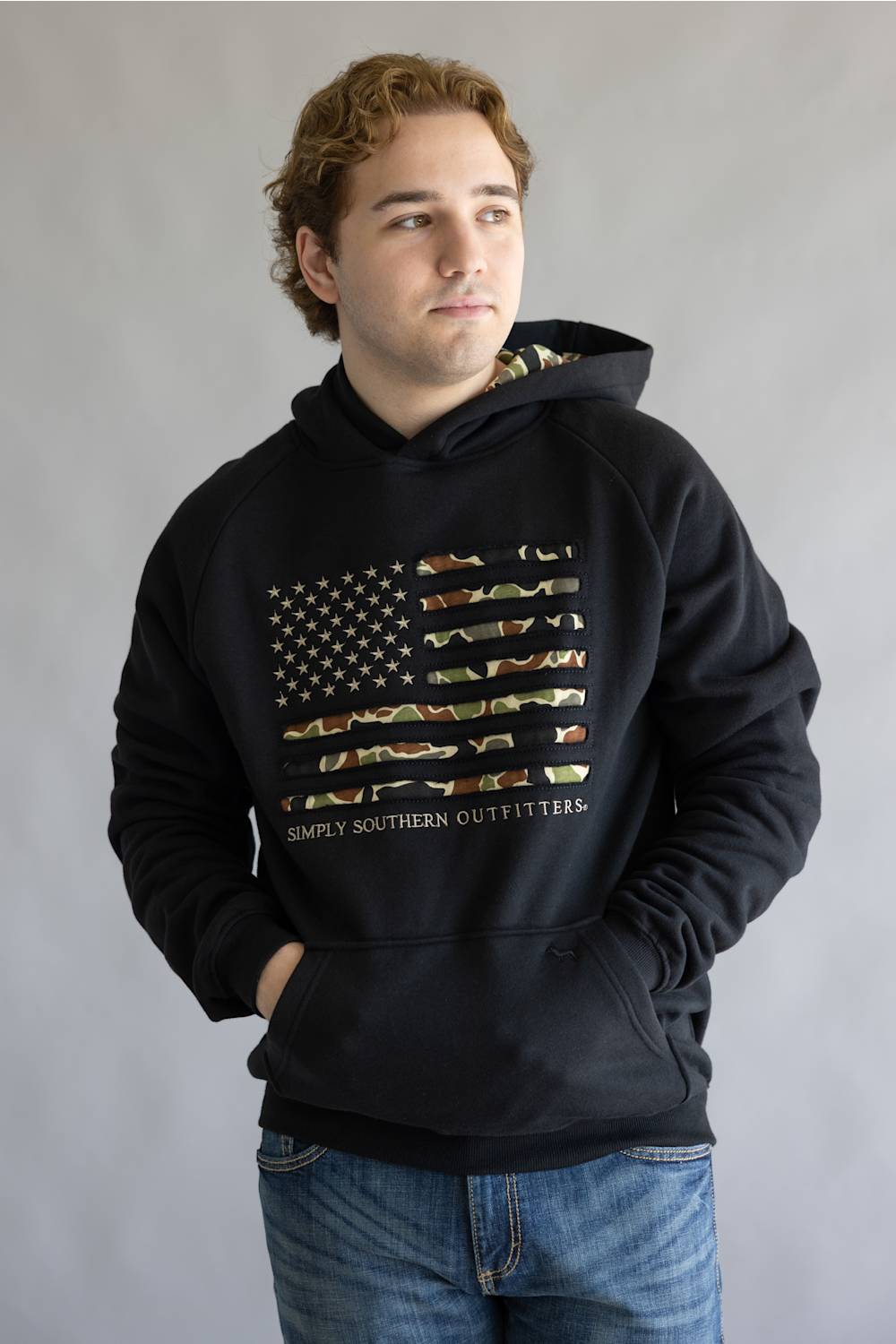 A man wearing a black pullover hoodie with a distressed American flag graphic made of camouflage-patterned stripes and the text 'SIMPLY SOUTHERN OUTFITTERS' printed below it. He is also wearing blue jeans, standing against a neutral gray studio background.