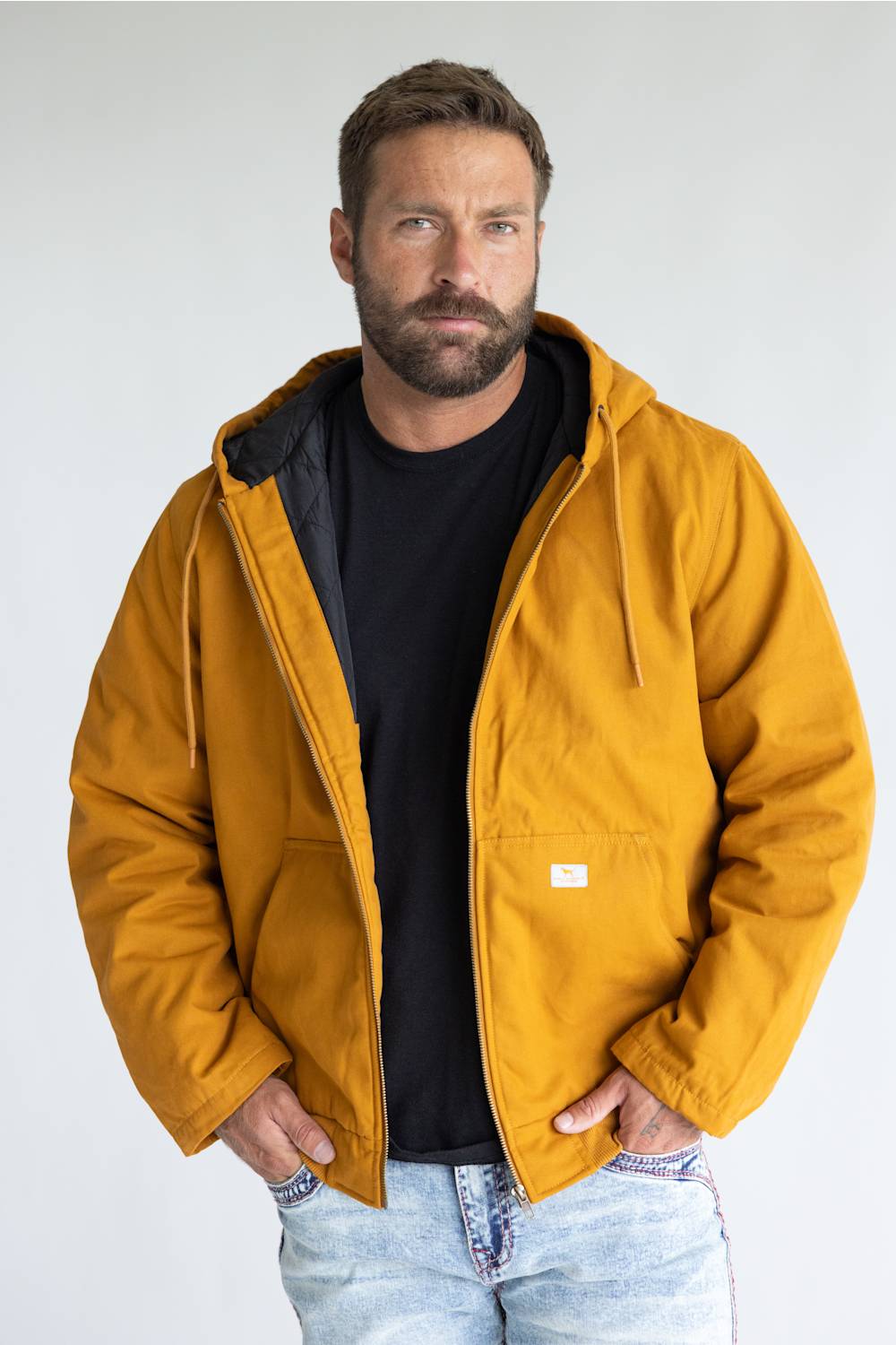 A man wearing a mustard yellow hooded work jacket over a black t-shirt and light blue jeans, standing against a light gray studio background.