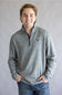 A smiling young man wearing a heather gray quarter-zip pullover sweater with a small black bird logo on the chest, paired with blue jeans, standing against a neutral gray studio background.