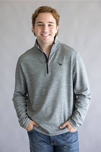 A smiling young man wearing a heather gray quarter-zip pullover sweater with a small black bird logo on the chest, paired with blue jeans, standing against a neutral gray studio background.