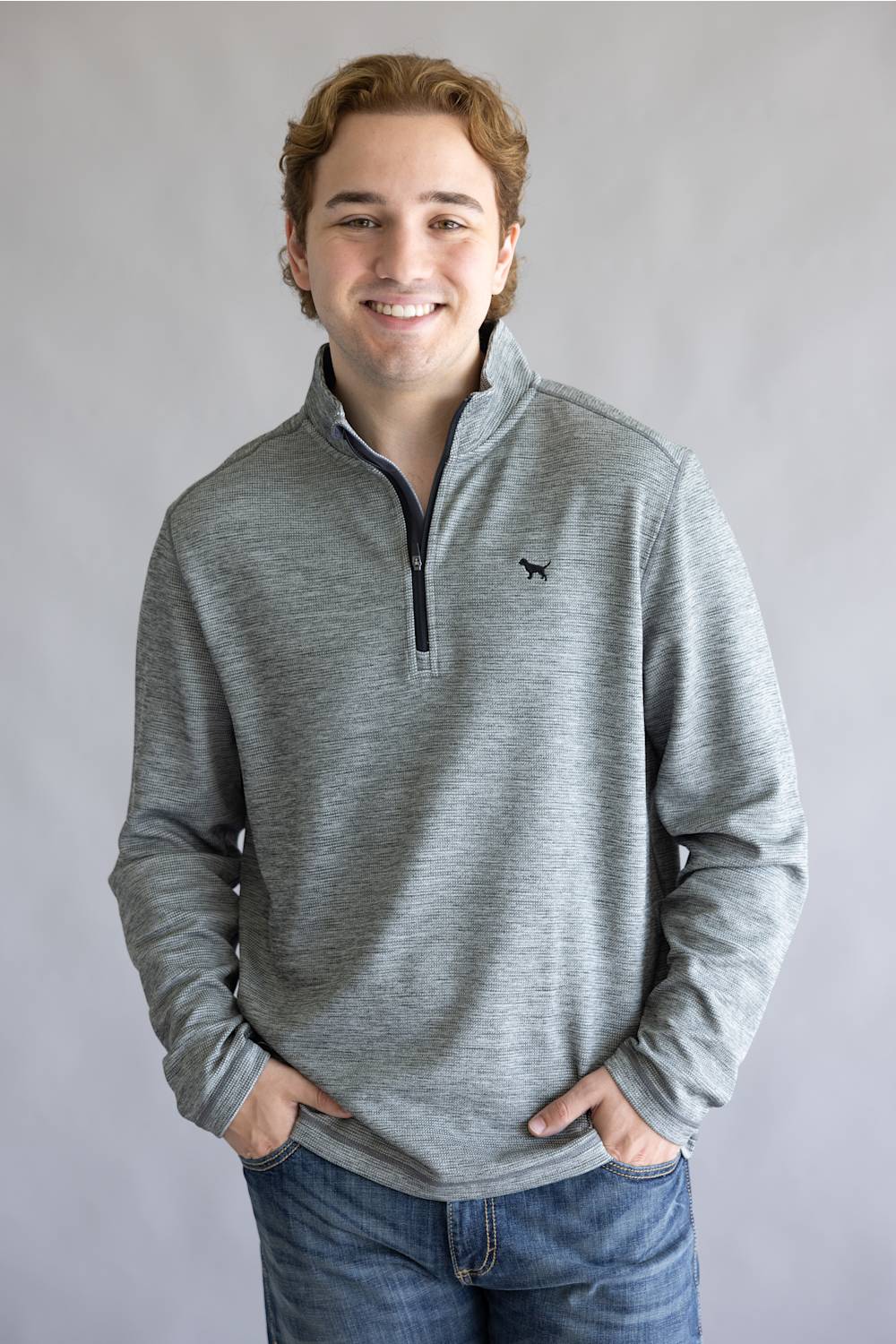 A smiling young man wearing a heather gray quarter-zip pullover sweater with a small black bird logo on the chest, paired with blue jeans, standing against a neutral gray studio background.