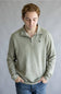 A man wearing a light sage green heathered quarter-zip pullover sweater with a small black dog logo on the left chest, paired with blue jeans, standing against a neutral gray background.