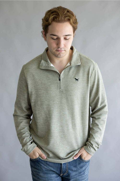 A man wearing a light sage green heathered quarter-zip pullover sweater with a small black dog logo on the left chest, paired with blue jeans, standing against a neutral gray background.