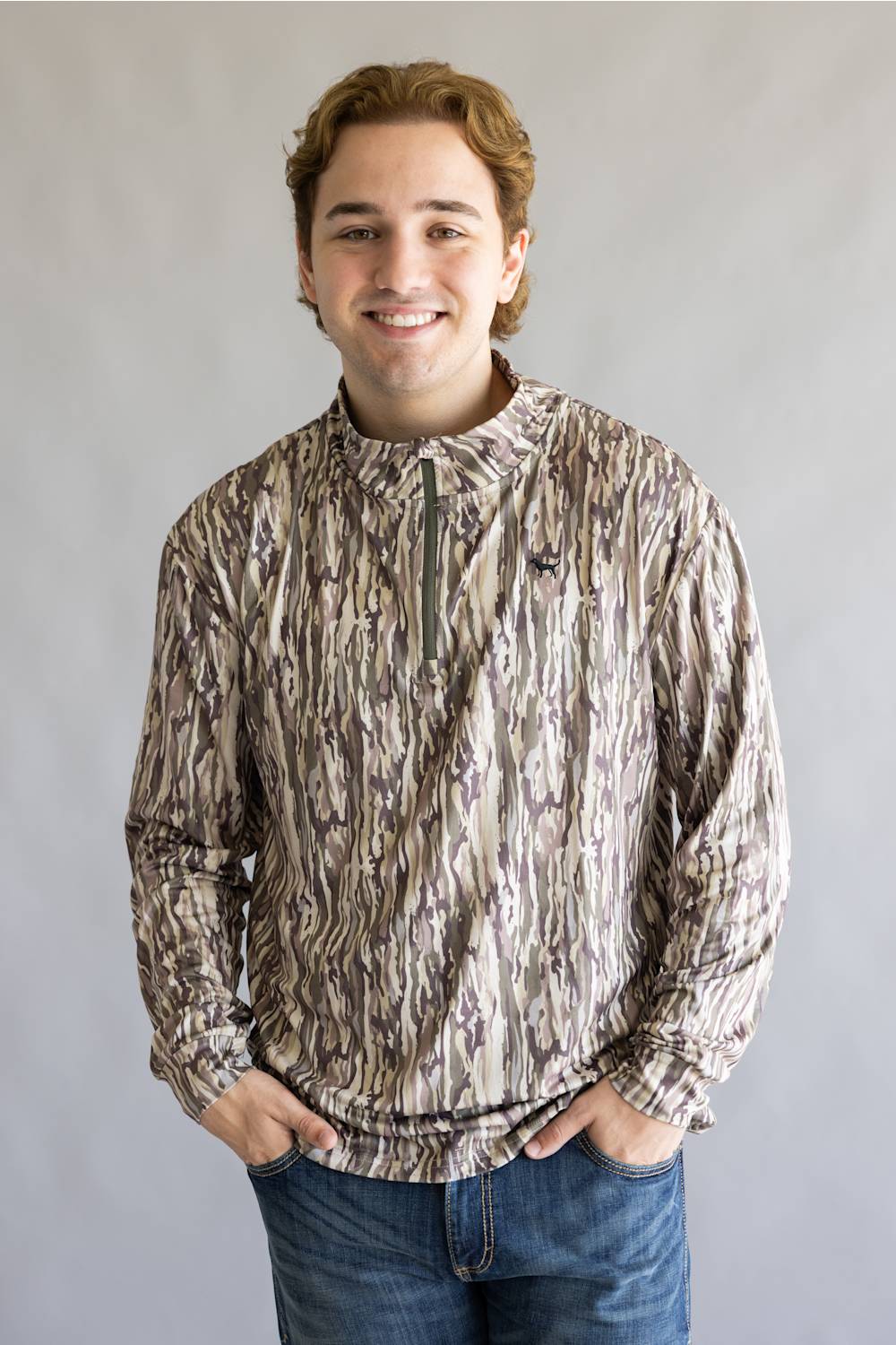 A smiling young man wearing a long-sleeve quarter-zip pullover with a brown, beige, and gray bark-like camouflage pattern, paired with blue jeans, standing against a neutral gray background.