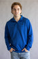 A young man with light brown hair wearing a textured royal blue quarter-zip pullover sweater and light-wash jeans, standing against a neutral gray studio background.