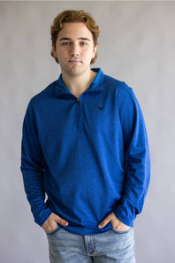 A young man with light brown hair wearing a textured royal blue quarter-zip pullover sweater and light-wash jeans, standing against a neutral gray studio background.