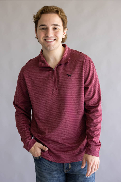 A smiling young man wearing a maroon long-sleeve quarter-zip pullover with a small black bird logo on the left chest, paired with blue jeans, standing against a neutral gray studio background.