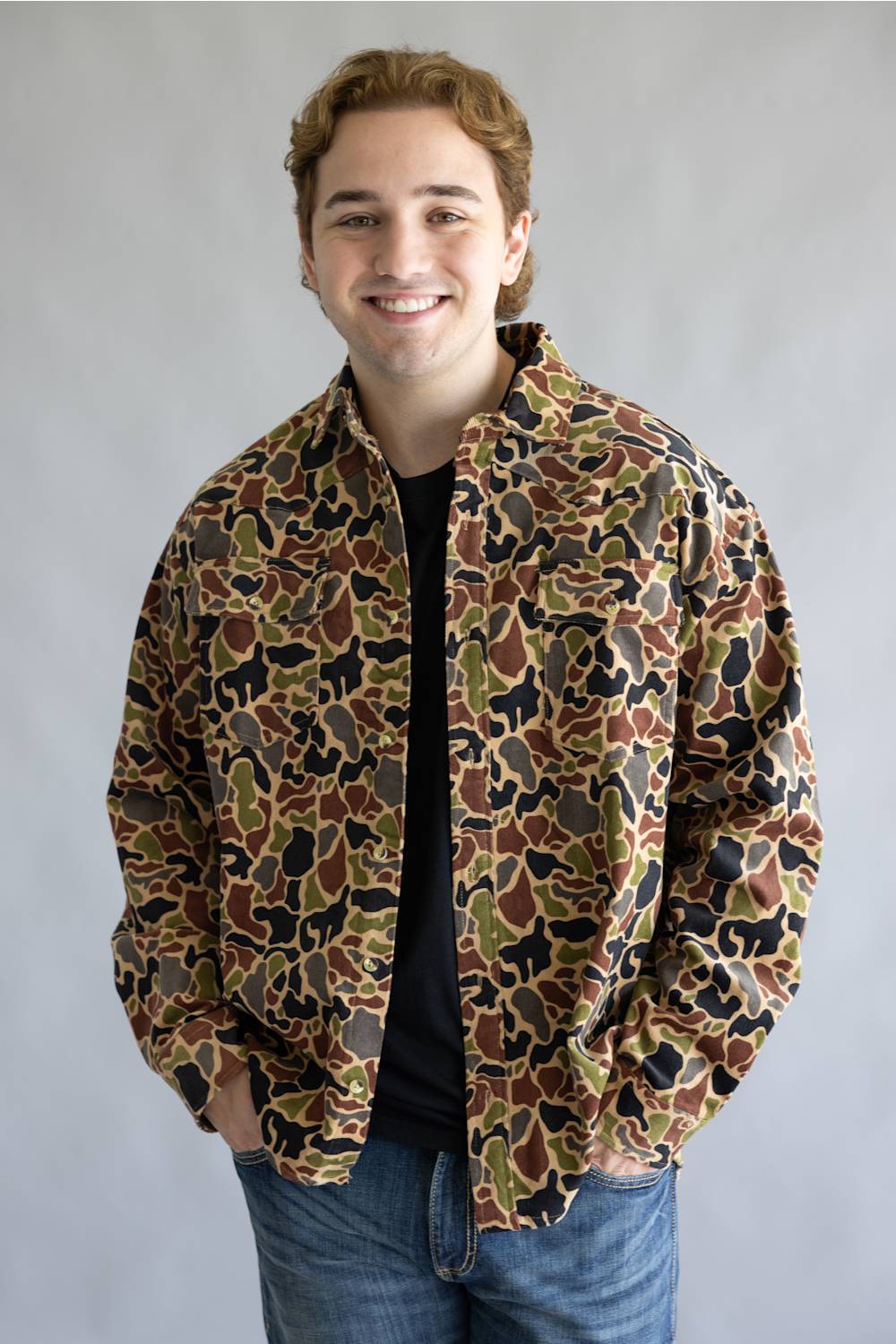 A smiling young man wearing a camouflage print jacket over a black t-shirt and blue jeans, standing against a plain gray studio background.