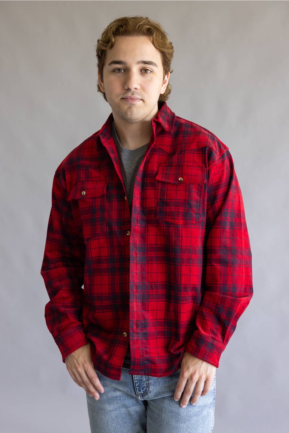 A young man wearing a red and black plaid flannel button-up shirt over a gray t-shirt and blue jeans, standing against a neutral gray studio background.