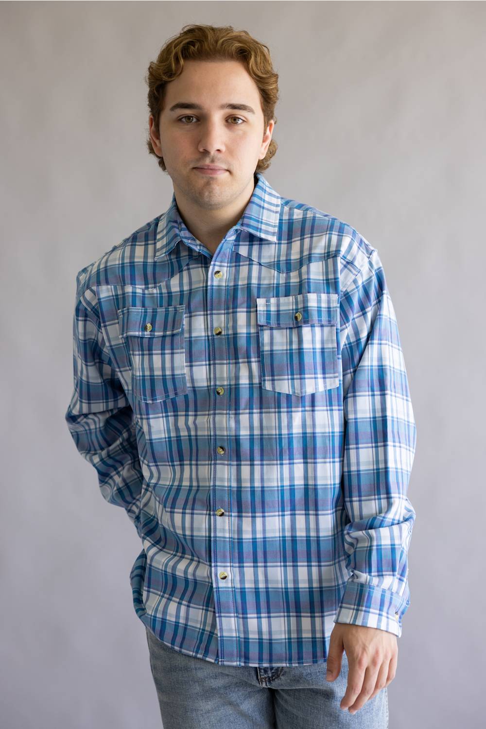 A man wearing a blue and white plaid long-sleeve button-up shirt with two chest pockets, paired with light blue jeans, standing against a neutral gray studio background.