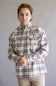 A man wearing a long-sleeve plaid button-down shirt in beige, brown, and white, paired with khaki pants, standing against a neutral gray studio background.