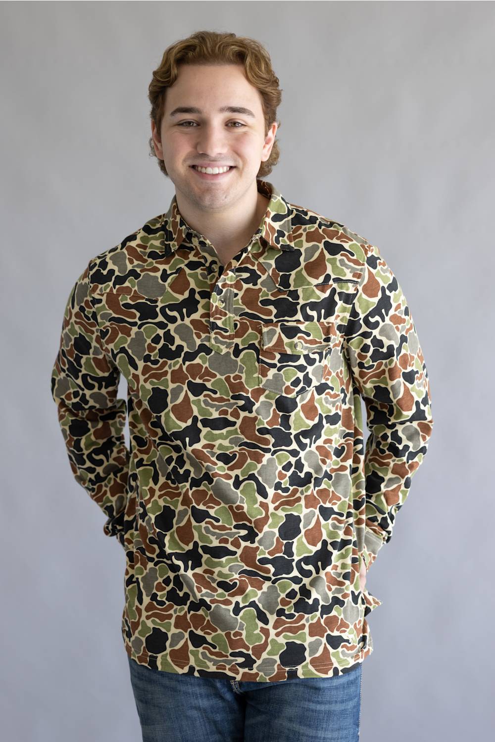 A smiling young man wearing a long-sleeve button-up shirt with an all-over camouflage pattern in brown, green, beige, and black, paired with blue jeans, set against a plain gray studio background.
