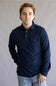 A man wearing a dark navy blue long-sleeve button-up shirt with a chest pocket, paired with light blue jeans, standing against a neutral gray studio background.