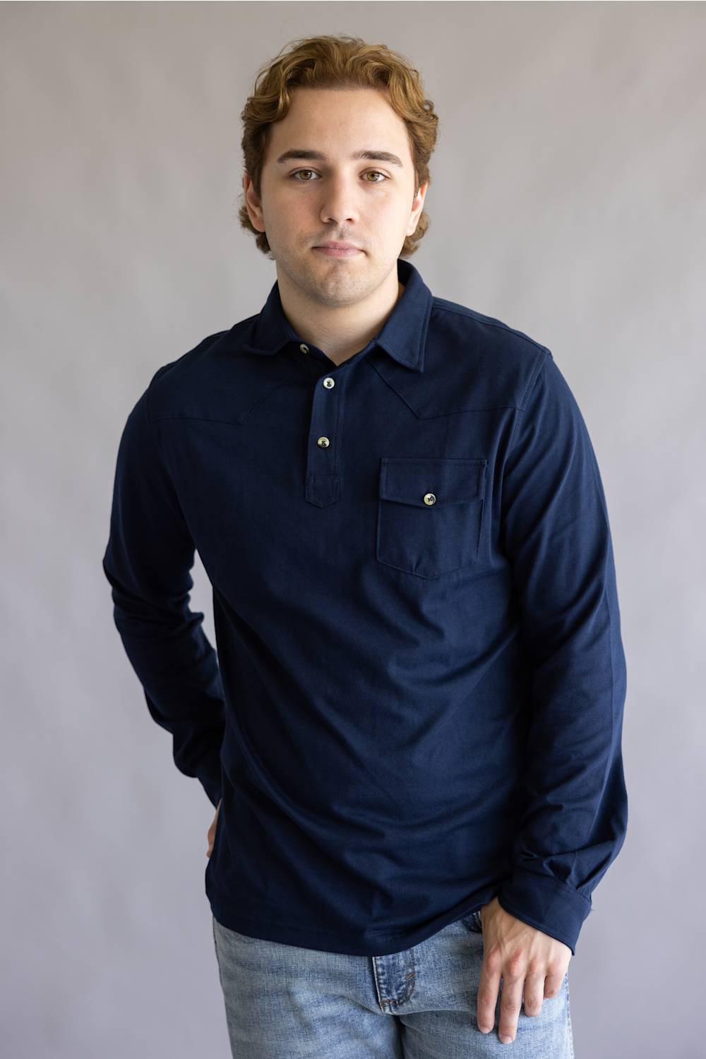 A man wearing a dark navy blue long-sleeve button-up shirt with a chest pocket, paired with light blue jeans, standing against a neutral gray studio background.