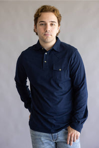 A man wearing a dark navy blue long-sleeve button-up shirt with a chest pocket, paired with light blue jeans, standing against a neutral gray studio background.