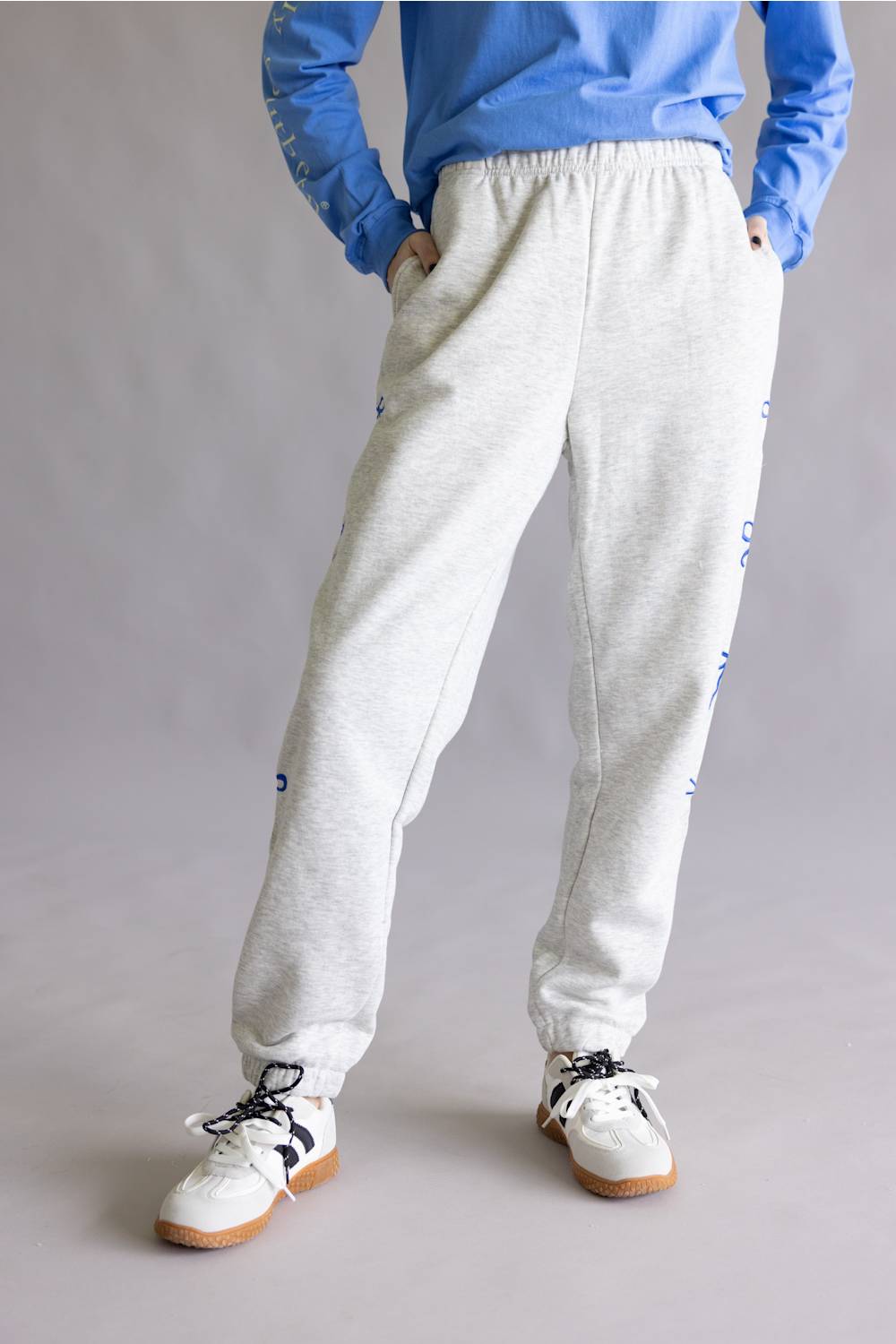 A person wearing light heather gray jogger sweatpants with blue side graphics, paired with a blue long-sleeve top and white sneakers, shown from the waist down against a plain gray studio background.