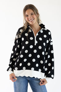 A woman modeling a black long-sleeve half-zip hoodie with large white polka dots and a white scalloped hem, paired with blue jeans against a plain white background.