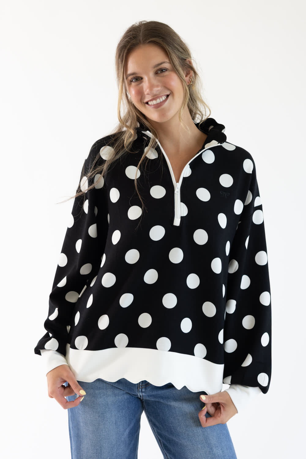 A woman modeling a black long-sleeve half-zip hoodie with large white polka dots and a white scalloped hem, paired with blue jeans against a plain white background.
