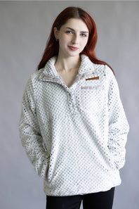 A woman with red hair wearing a white textured fleece pullover with a small brown logo patch on the chest, photographed against a neutral gray studio background.