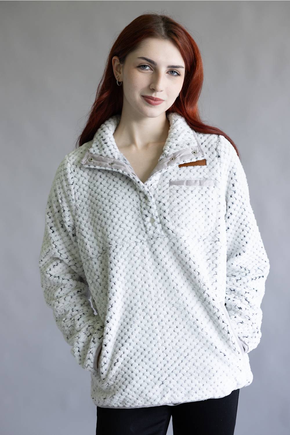 A woman with red hair wearing a white textured fleece pullover with a small brown logo patch on the chest, photographed against a neutral gray studio background.