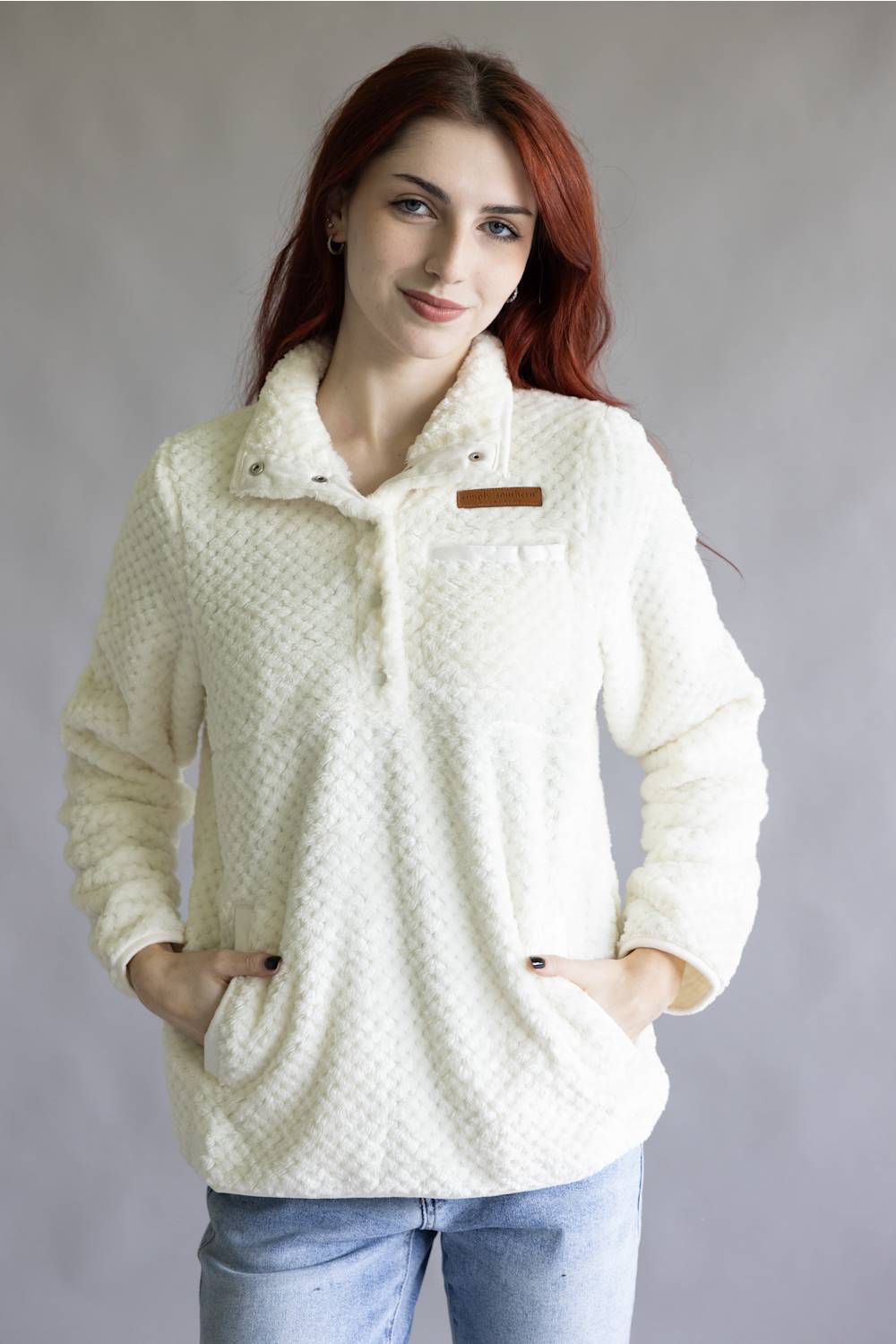 A woman with red hair wearing a cream-colored textured fleece pullover jacket with a stand-up collar, snap buttons, and a chest pocket with a brown leather accent, paired with light blue jeans, against a plain gray studio background.