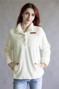 A woman with red hair wearing a cream-colored textured fleece pullover jacket with a stand-up collar, snap buttons, and a chest pocket with a brown leather accent, paired with light blue jeans, against a plain gray studio background.