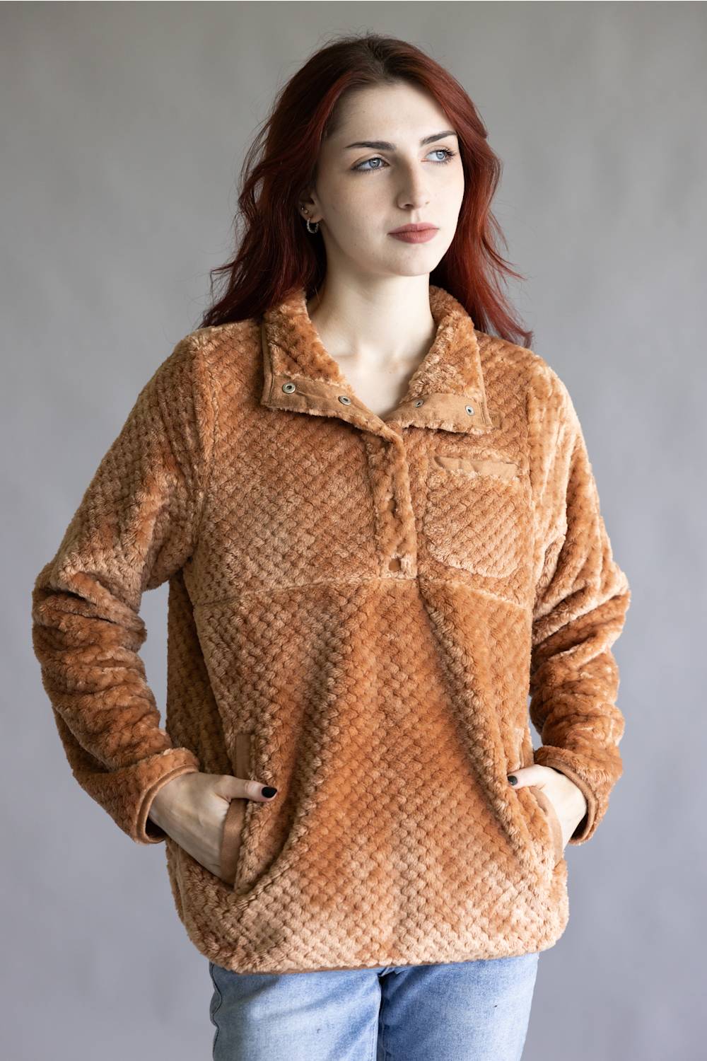A woman with red hair wearing a textured, warm brown fleece pullover jacket with a buttoned collar and front pockets, paired with blue jeans, against a plain gray studio background.