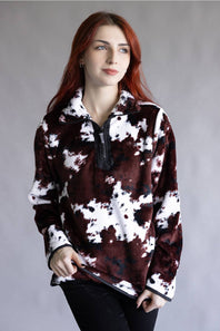 A woman with red hair wearing a brown and white cow-print fleece quarter-zip pullover, paired with black pants, standing against a neutral gray studio background.