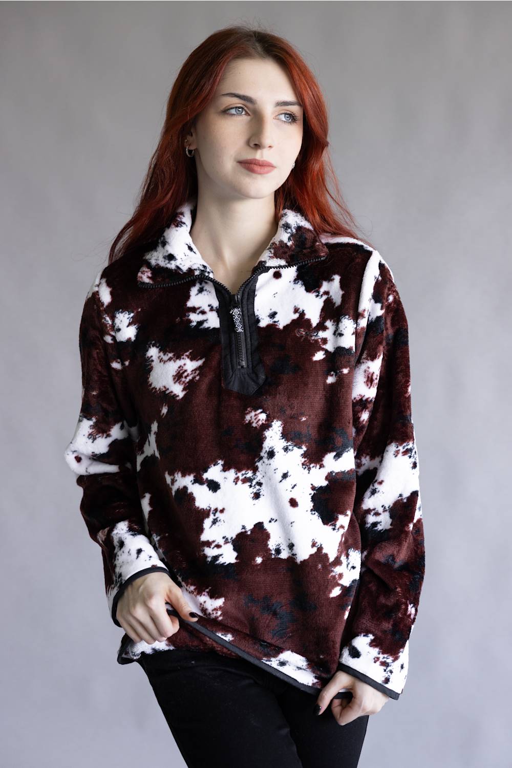 A woman with red hair wearing a brown and white cow-print fleece quarter-zip pullover, paired with black pants, standing against a neutral gray studio background.