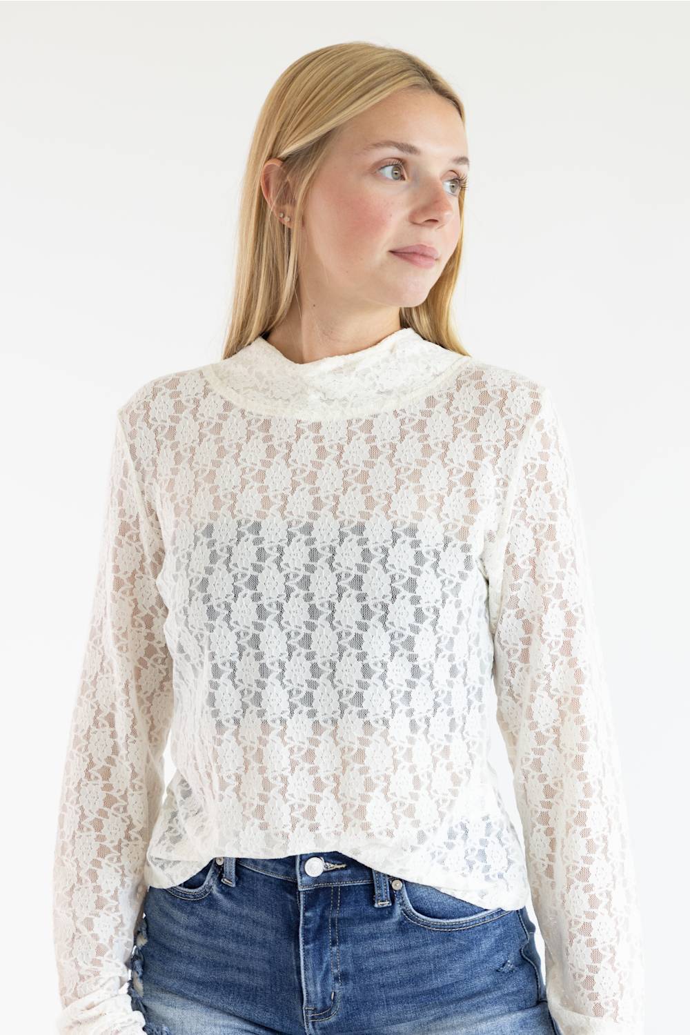 A woman wearing a white lace mock-neck long-sleeve top and blue jeans, photographed against a plain white studio background.