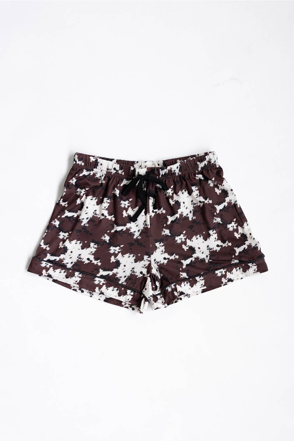 A pair of youth girls shorts featuring a dark brown and white cow pattern, with a black drawstring at the elastic waistband, laid flat against a white background.