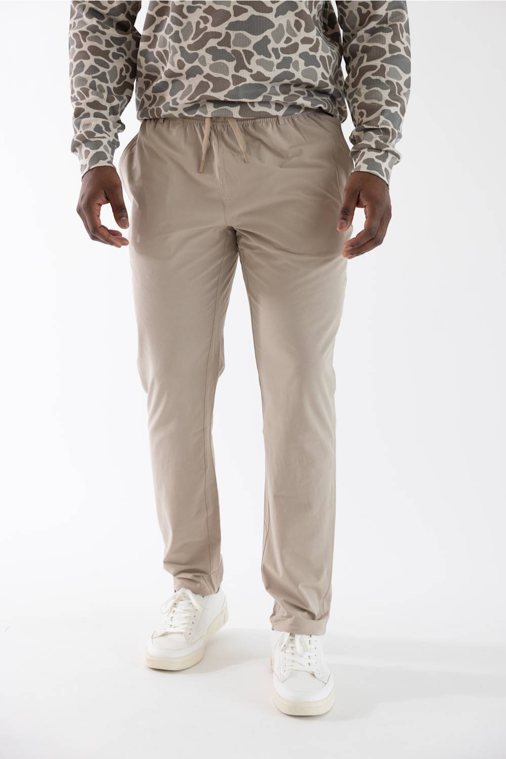 A person wearing beige drawstring jogger pants, a camouflage-patterned long-sleeve shirt, and white sneakers, photographed against a clean white studio background.