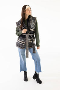 A woman modeling a sleeveless black and white patterned knit vest with a matching tie belt, worn over an olive green long-sleeve top, blue jeans, and black ankle boots, against a plain white studio background.