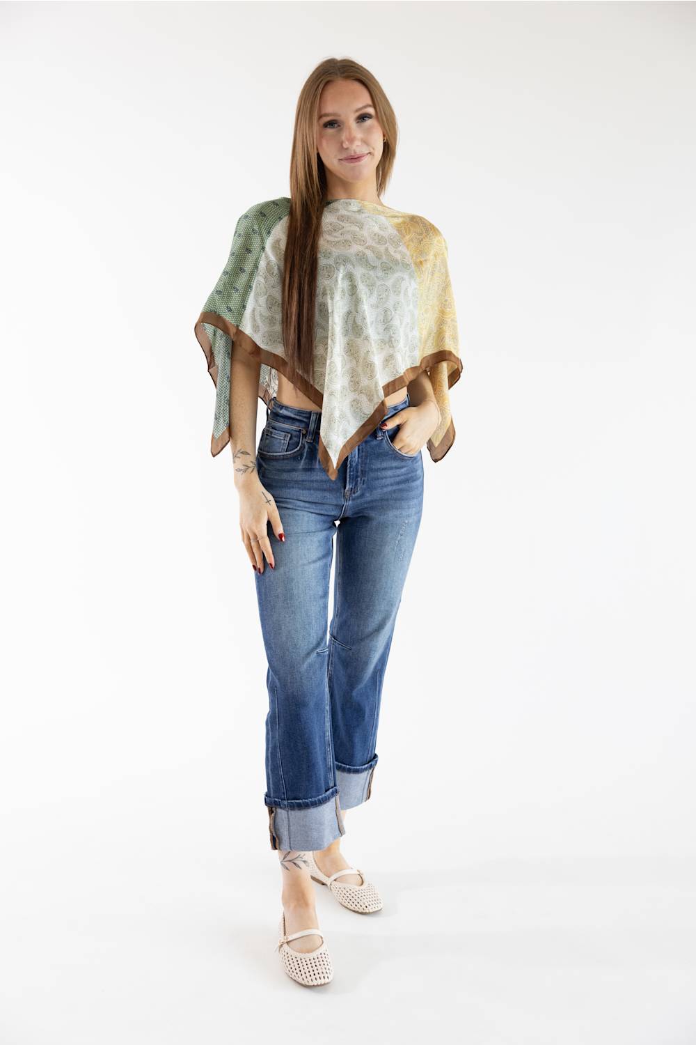 A woman modeling a lightweight poncho-style top with a multicolor abstract print in sage green, cream, and tan, paired with blue jeans and beige flat sandals, against a plain white studio background.