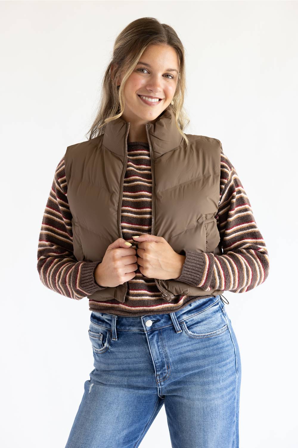 A woman wearing a brown puffer vest over a brown and red striped knit sweater, paired with blue jeans, photographed against a plain white studio background.