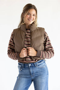 A woman wearing a brown puffer vest over a brown and red striped knit sweater, paired with blue jeans, photographed against a plain white studio background.