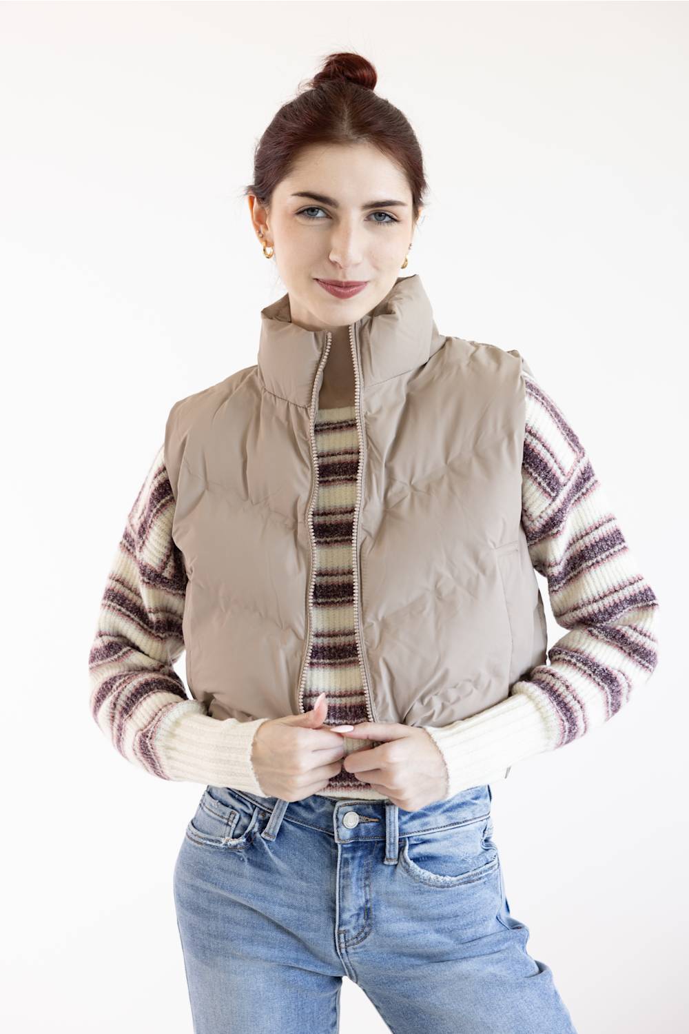 A woman wearing a beige puffer vest over a striped knit sweater and blue jeans, standing against a plain white studio background.